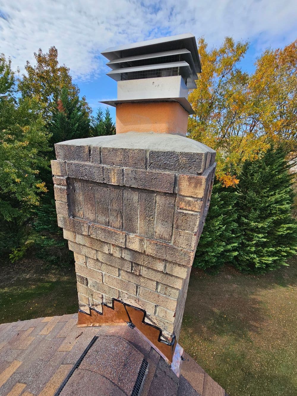 Chimney with stonework and metal cap against a backdrop of autumn trees and cloudy sky.