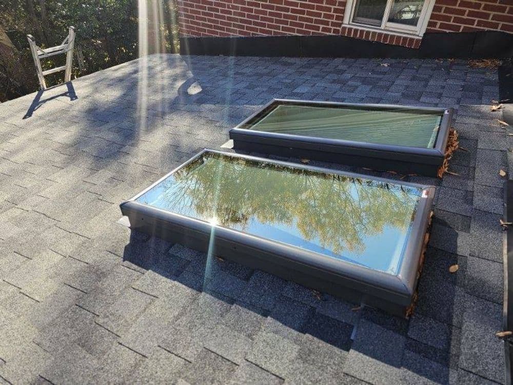 Two skylights on a shingled roof reflecting sunlight and trees, with a chair nearby.