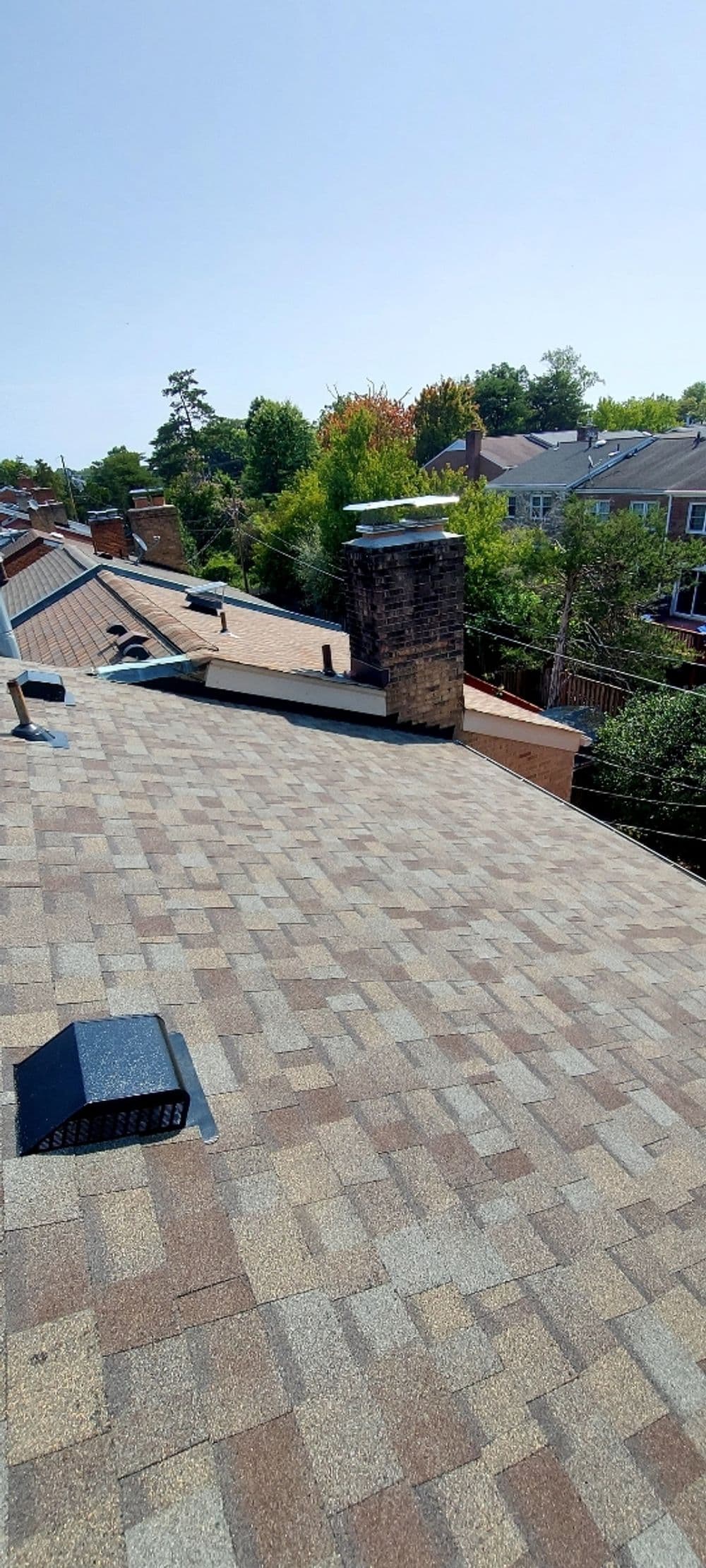 View of a residential roof with shingles and chimney, surrounded by trees and houses.