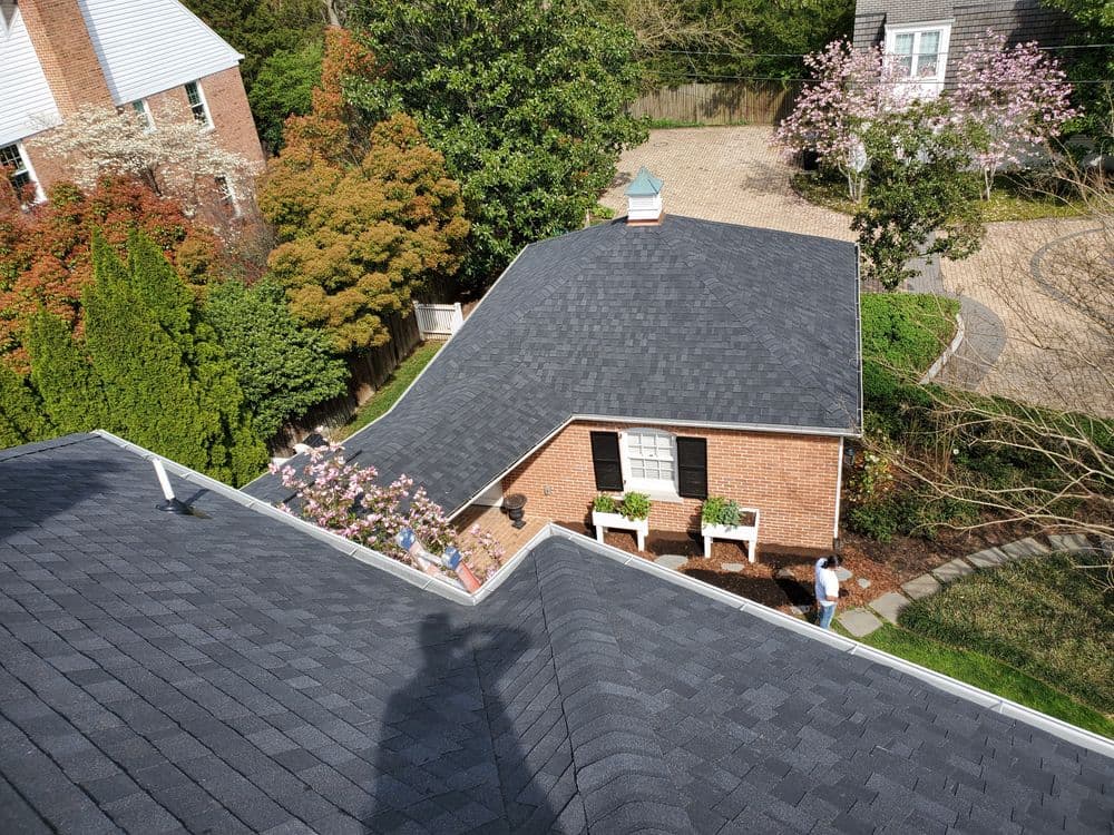 Aerial view of a residential roof with dark shingles, landscaped garden, and blooming trees.