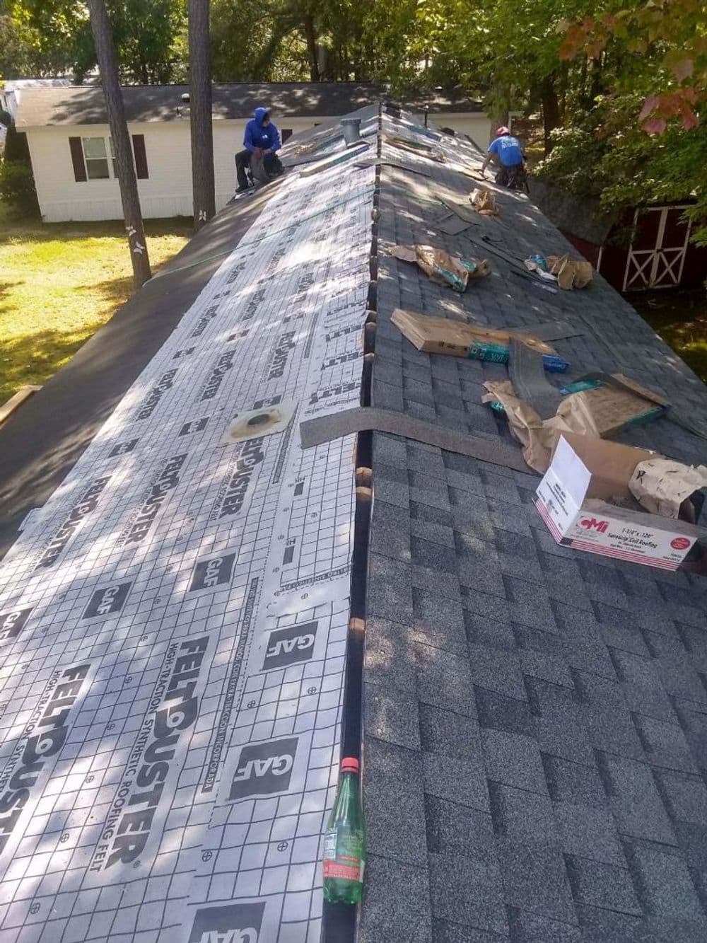 Workers installing roofing materials on a sloped roof surrounded by trees and houses.