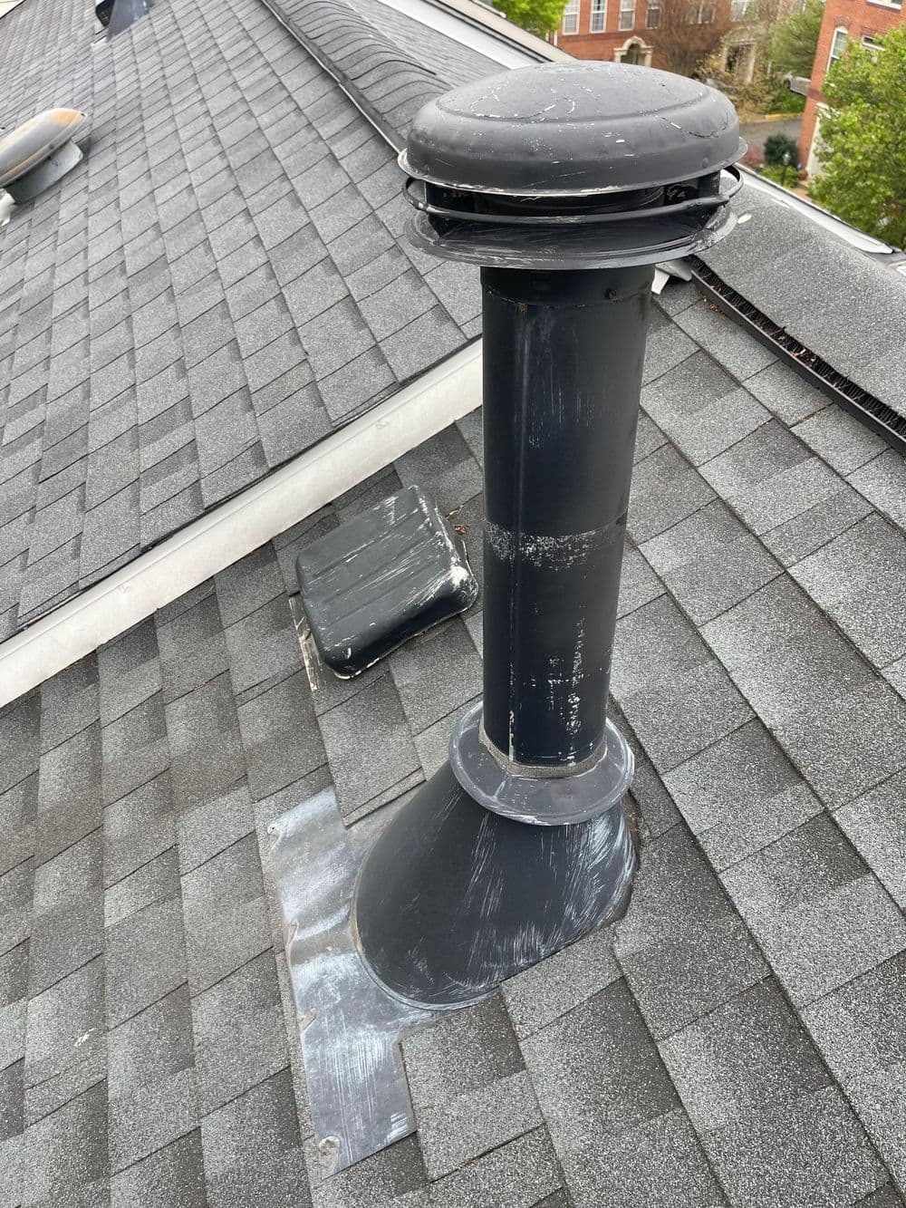 Black chimney vent pipe on a rooftop with shingles and surrounding vents.