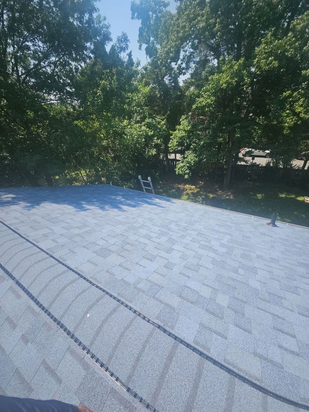 Aerial view of a newly shingled roof with trees and a ladder in the background.