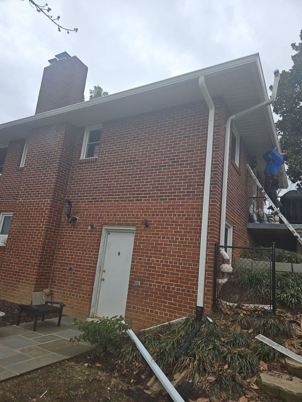 Worker performing exterior maintenance on a two-story brick house during overcast weather.