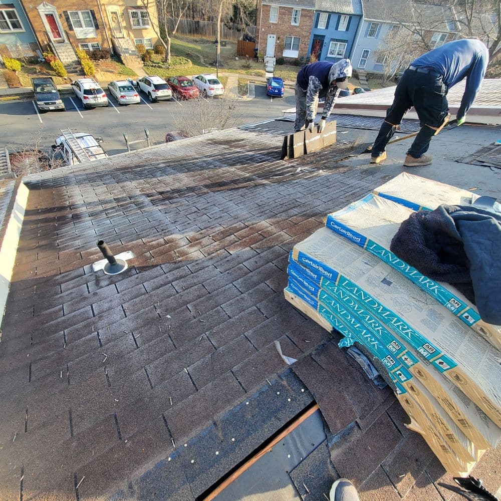 Roofing contractors installing shingles on a residential roof with shingles stacked nearby.