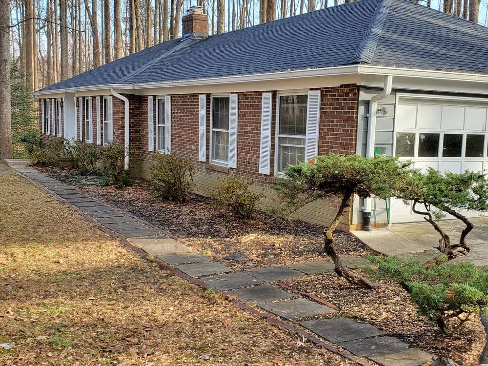 Charming brick ranch home surrounded by trees and landscaped garden, featuring a single car garage.