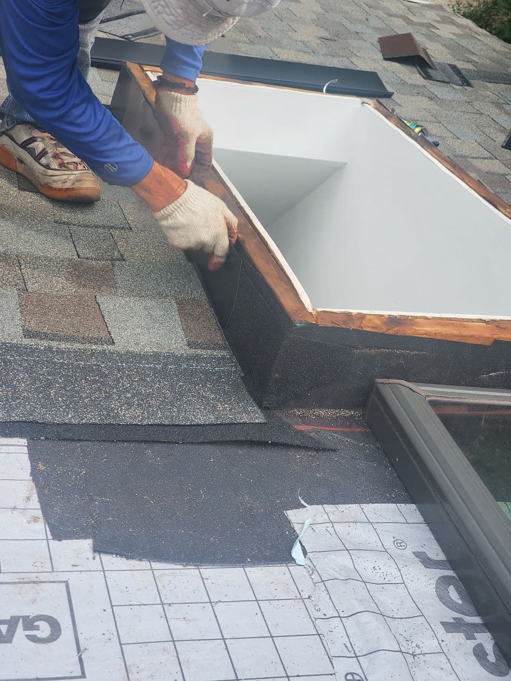 Worker installing a skylight on a shingled roof with tools and materials nearby.